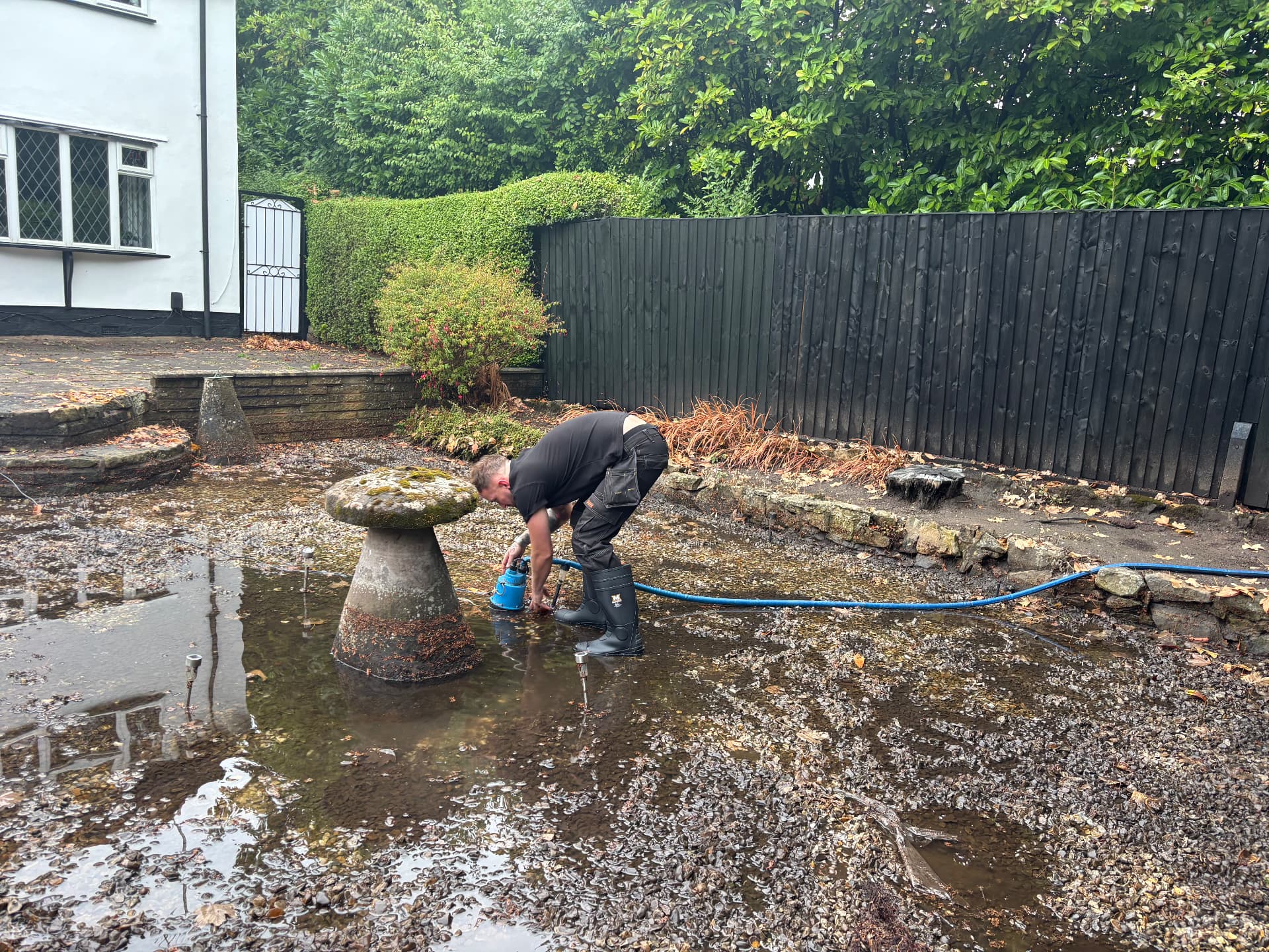 Technician positioning a submersible pump within standing water in a flooded gravelled garden
