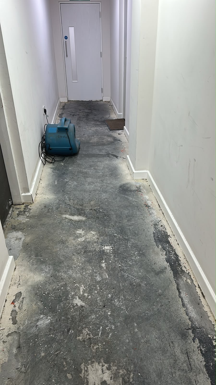 Stripped communal corridor with exposed damp screed and an air mover positioned against the wall