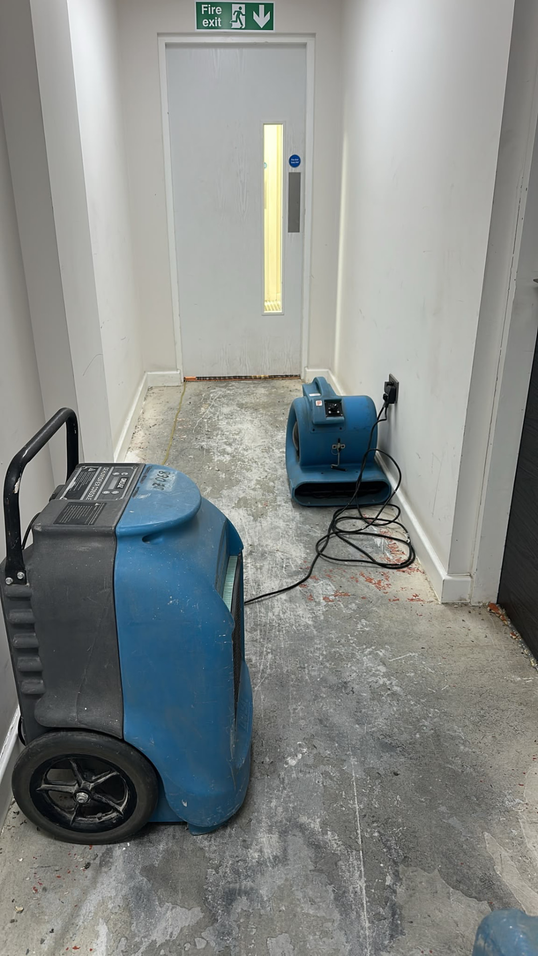 Dehumidifier and air mover running in a communal corridor beside a fire exit door